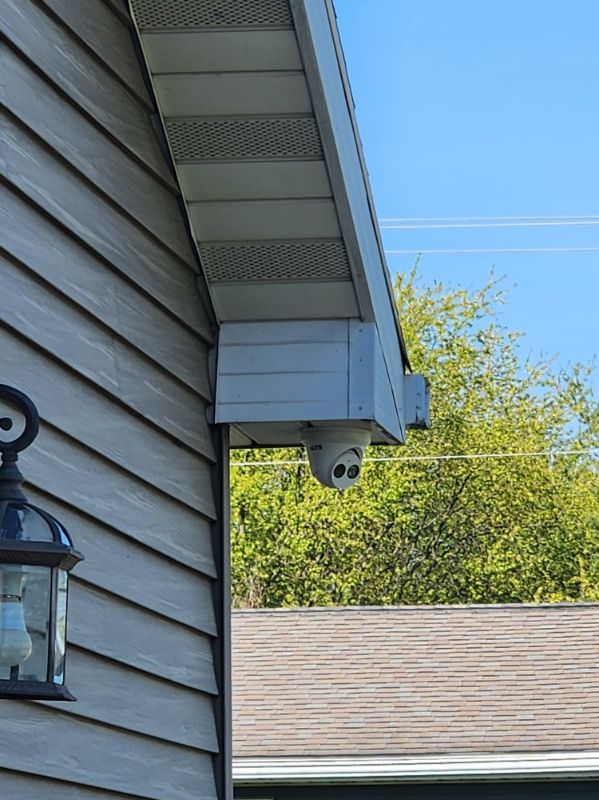 Exterior house corner with a mounted security camera under the eaves beside a lantern and siding