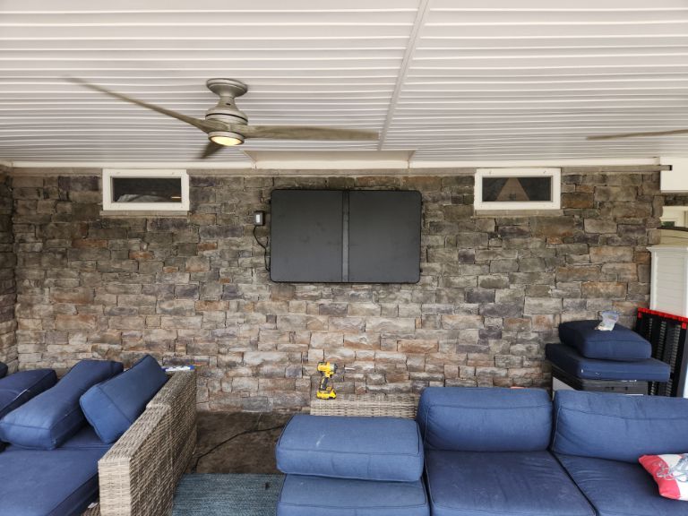 Covered patio with stone wall, blue wicker seating, ceiling fan, and wall-mounted TV.