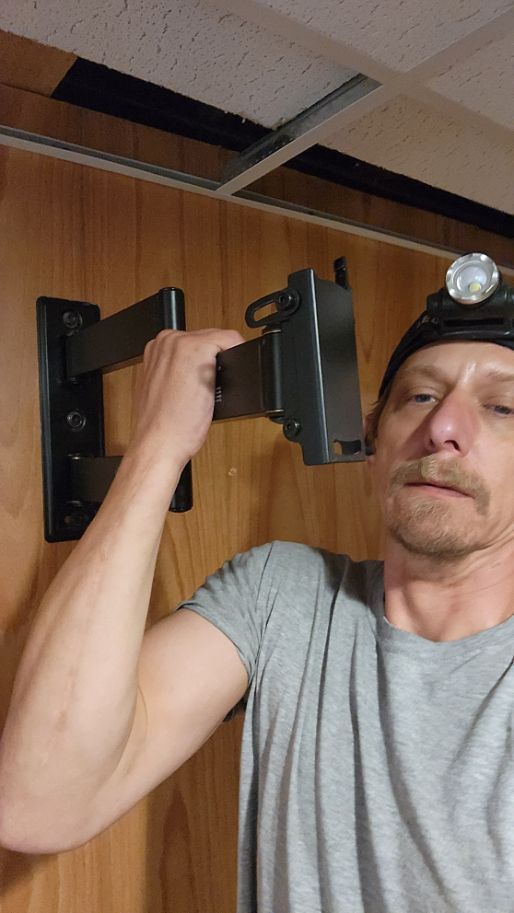 Man installing a wall-mounted TV bracket in a wood-paneled room, wearing a headlamp.