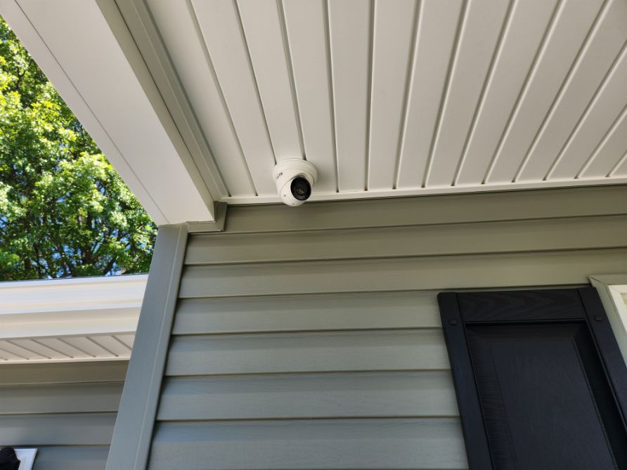 Outdoor security camera under a white roof overhang above gray siding and a window.