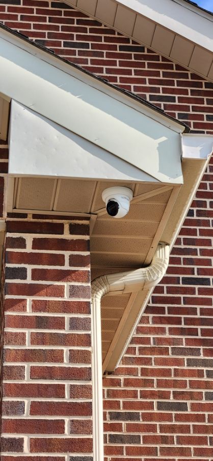 Security camera mounted under a brick house eave beside a white soffit and downspout