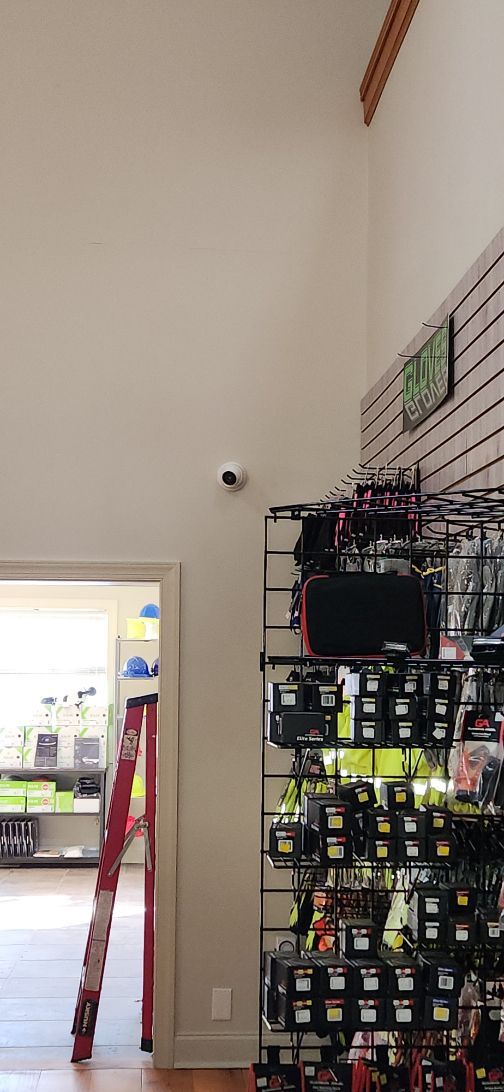 Store aisle with a tall white wall, security camera, and stacked black shopping baskets on shelving.