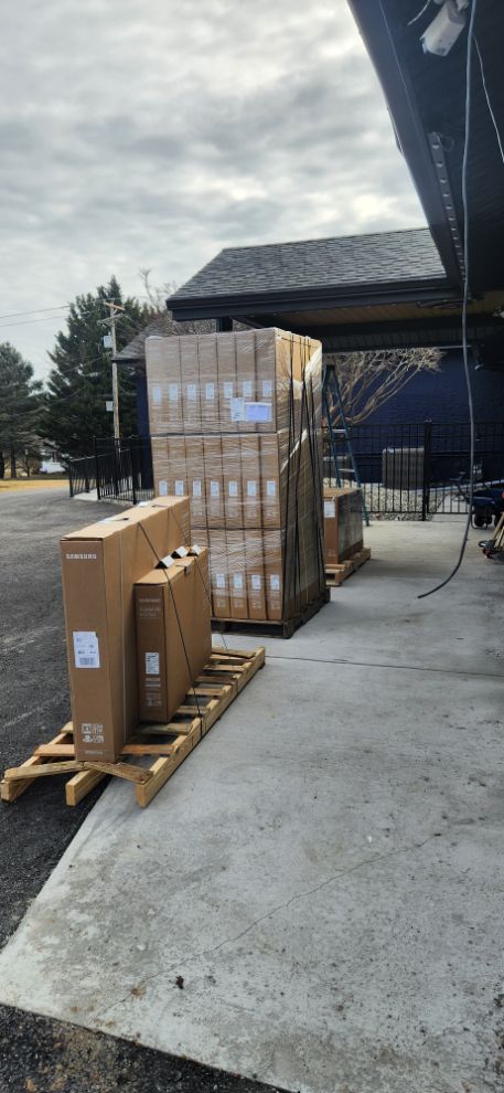 Pallets of boxed goods stacked outdoors beside a warehouse under an overcast sky