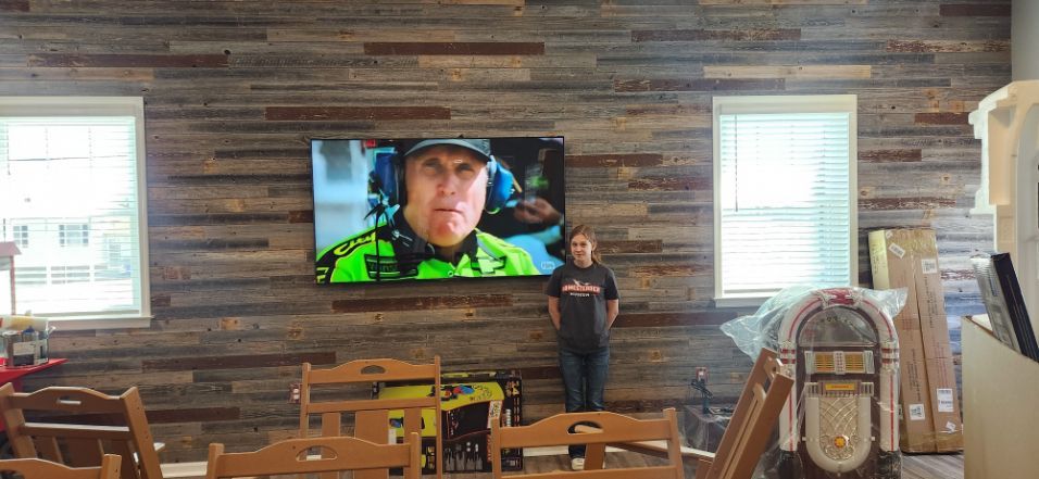 Person standing in a rustic room watching a wall-mounted TV above wooden tables and chairs
