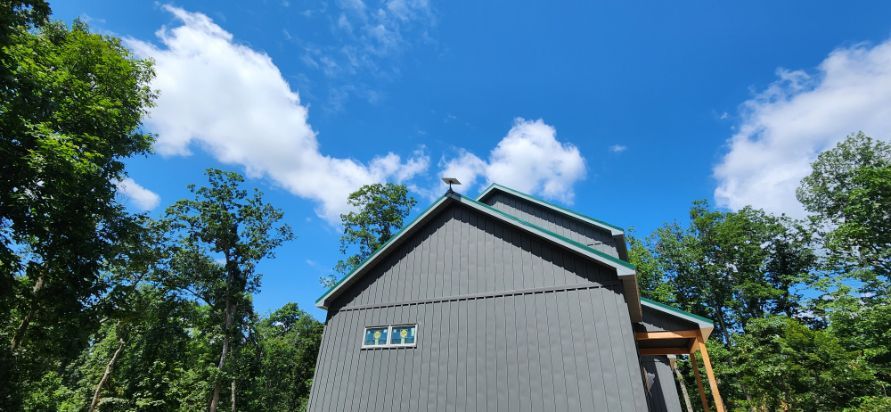 Gray house with a green roof under a bright blue sky, surrounded by trees