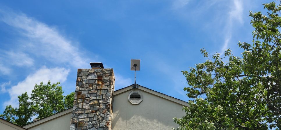 House roof with stone chimney beneath a blue sky, framed by green trees