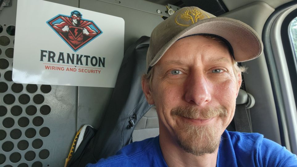 Man smiling in a vehicle cab beside a Frankton sign and license plate holder