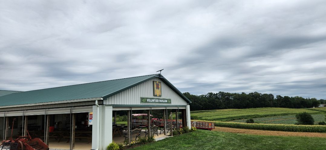 Farm stand with green roof beside rows of crops under a cloudy sky.