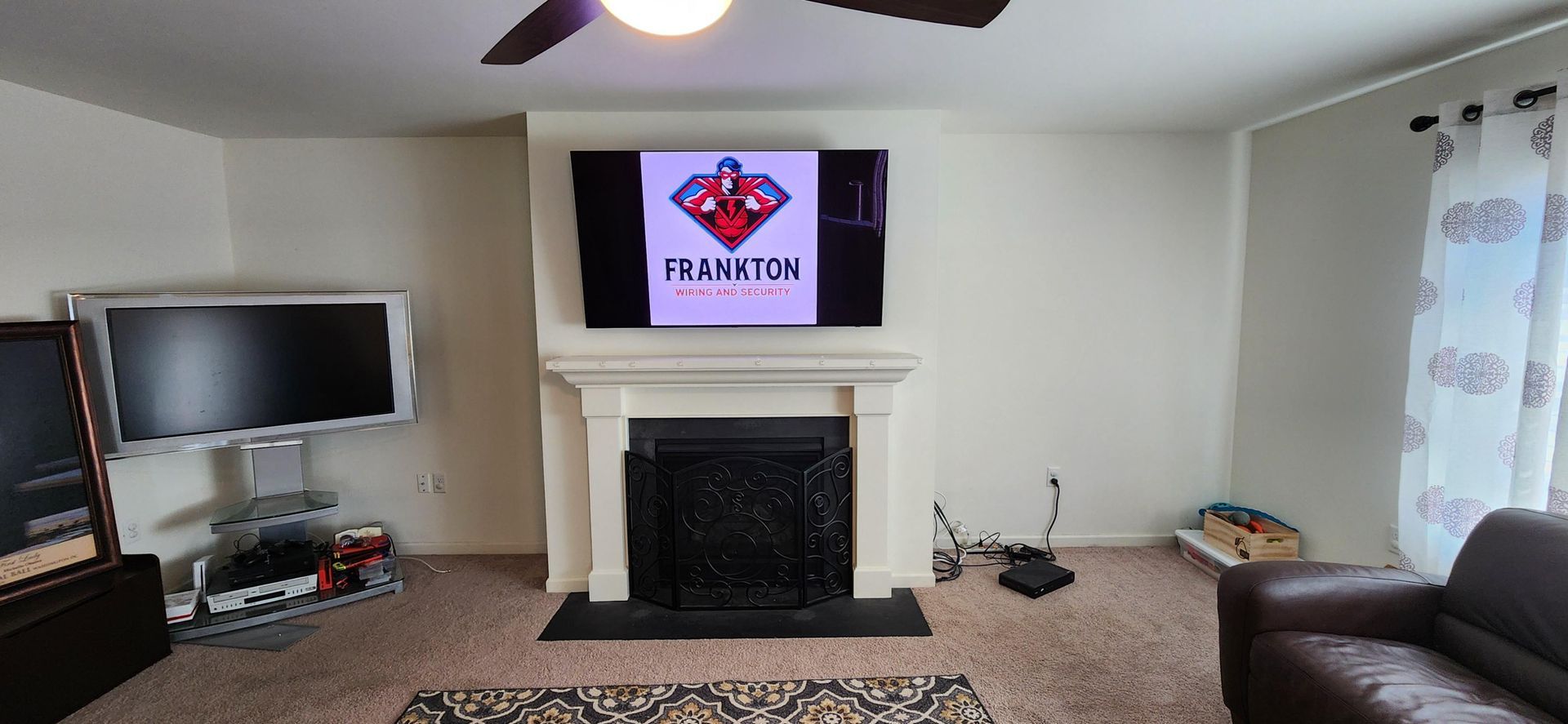 Bright living room with fireplace, TV, ceiling fan, patterned curtains, and dark sofa.