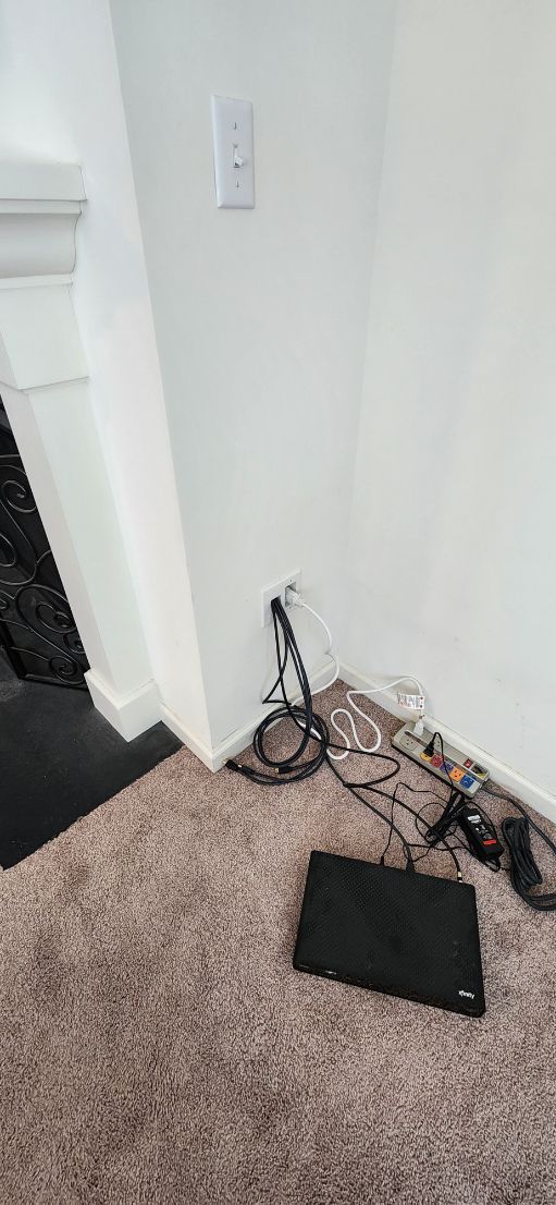 White wall corner with tangled cables, wall plate, carpet, and a black device on the floor near an outlet.