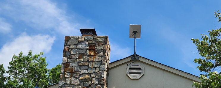 Stone chimney and house roof with a weather vane against a blue sky, framed by trees