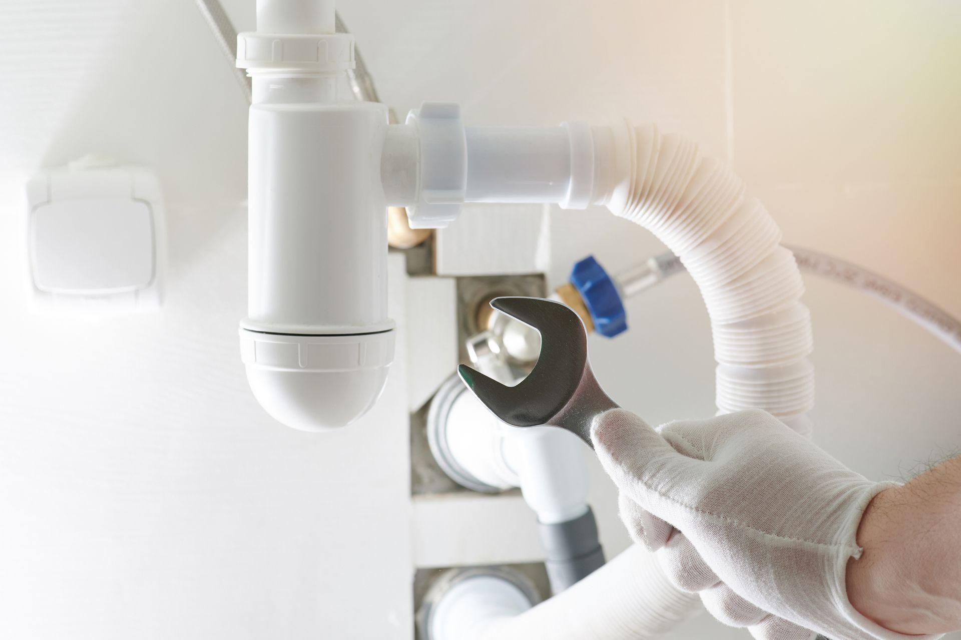 Plumber in white glove uses wrench to work on white sink pipes, under sink.