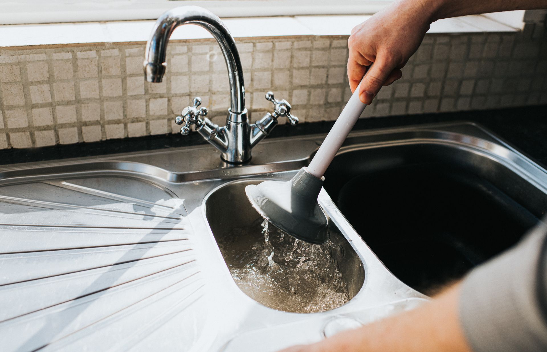 Person using a plunger in a stainless steel kitchen sink to unclog it.