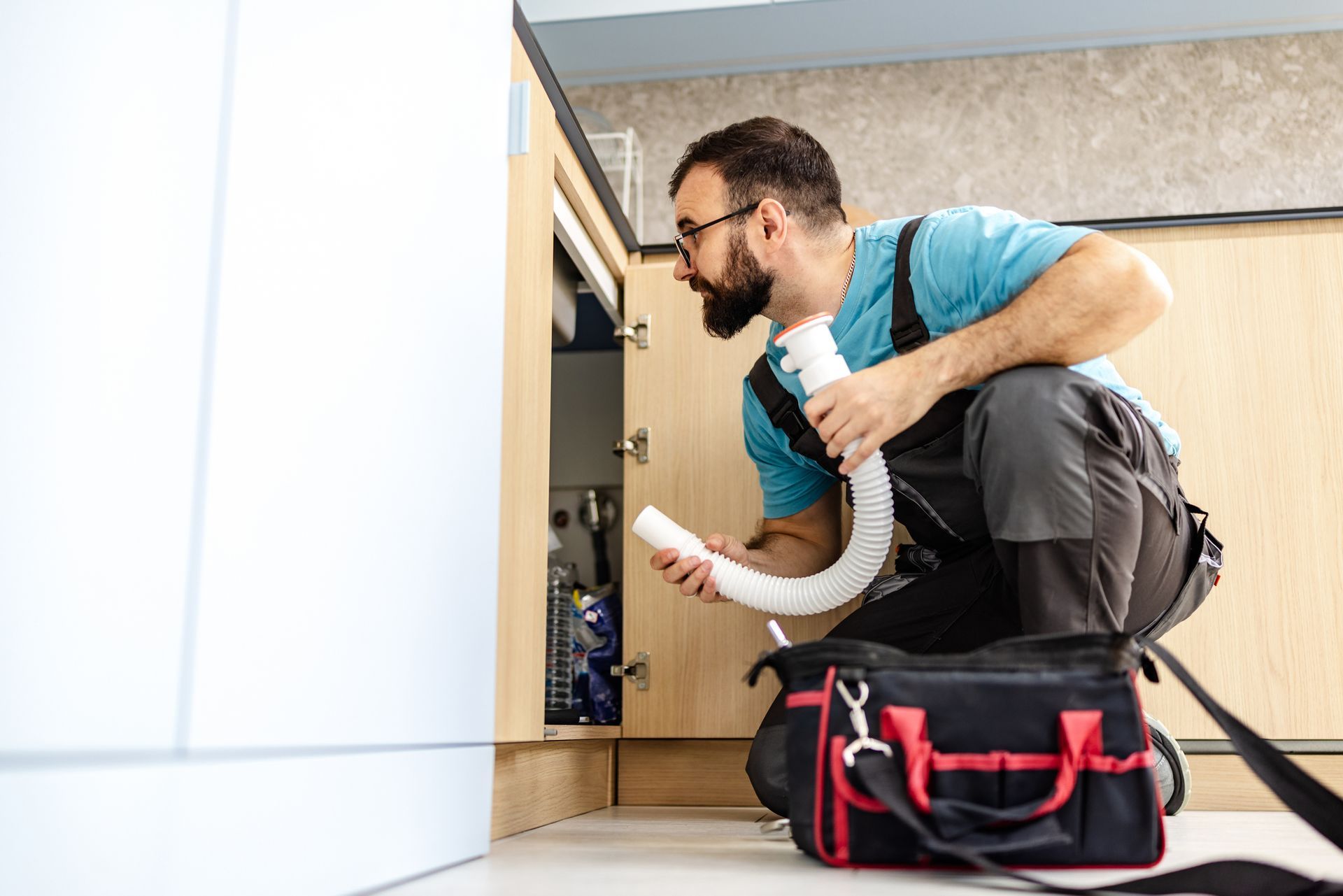 Plumber inspecting pipes under kitchen sink with toolbox.