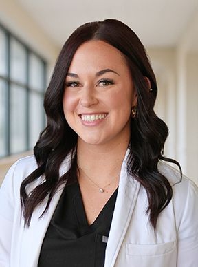 A woman in a white lab coat is smiling in front of a window.