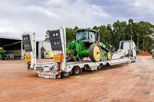Green tractor loaded on a white transport trailer, parked on red dirt.
