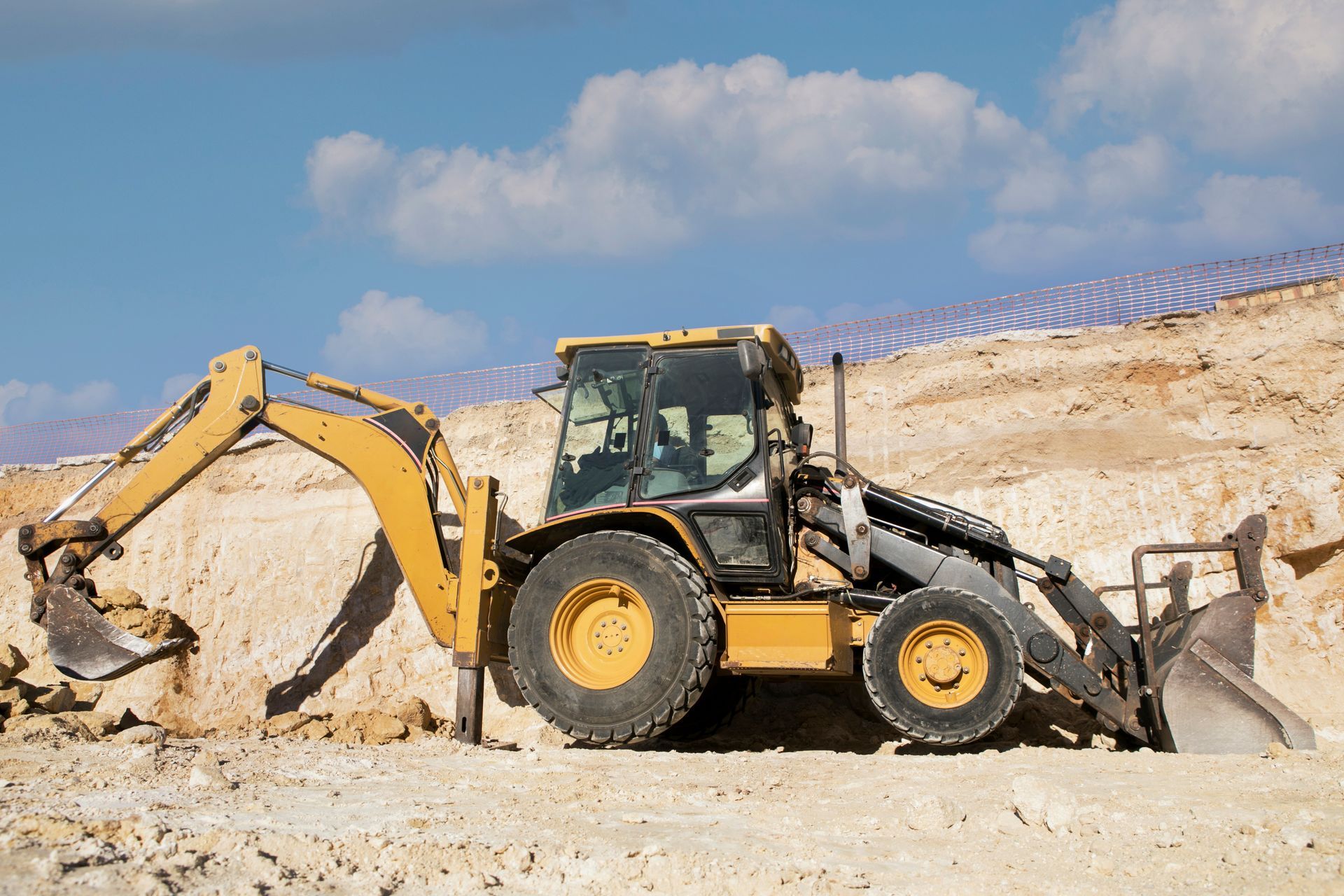 Yellow backhoe digging into a sandy hillside under a blue sky with clouds.
