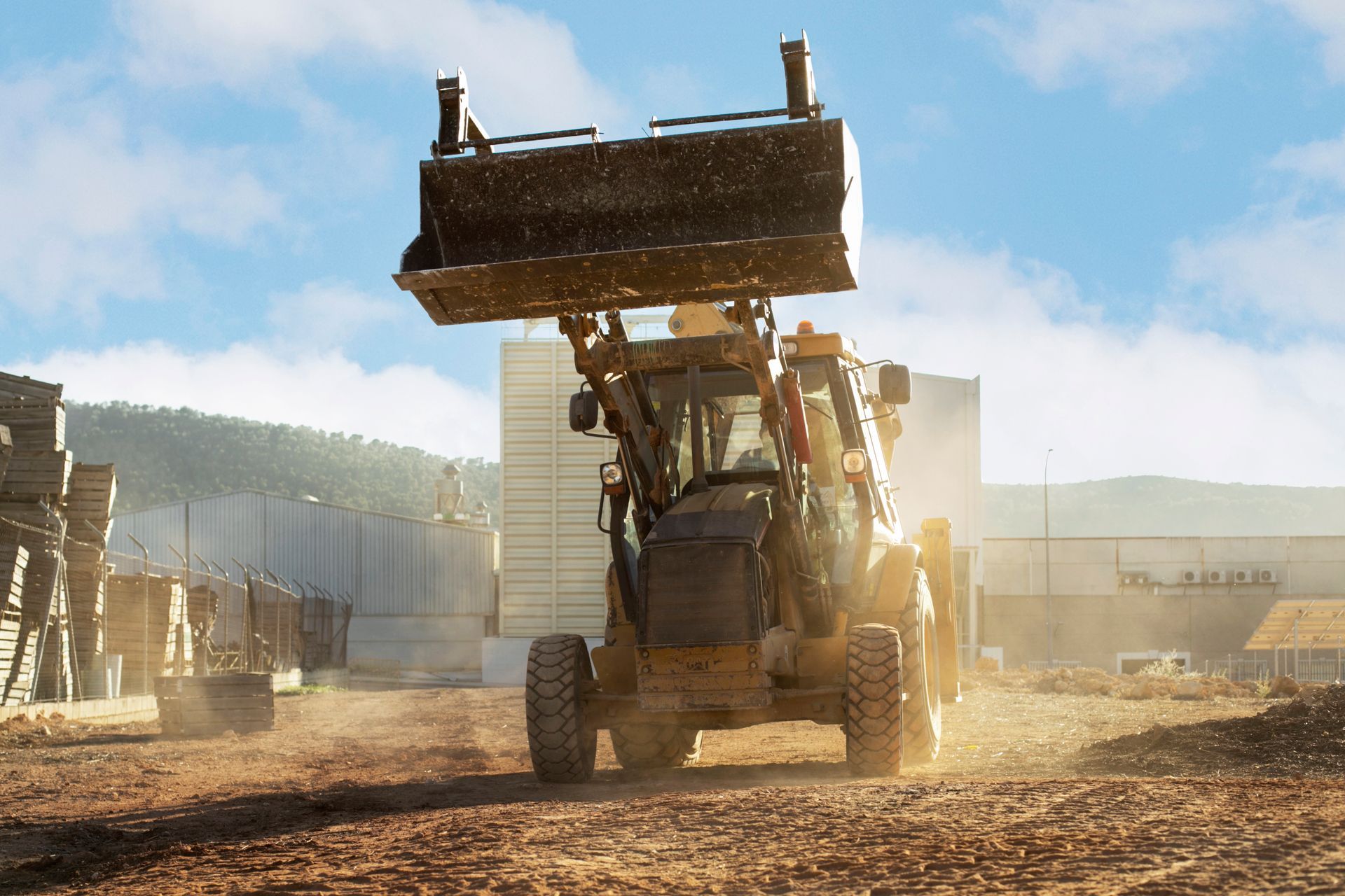 Yellow backhoe raises its bucket in a construction area, sunlit with a blue sky in the background.