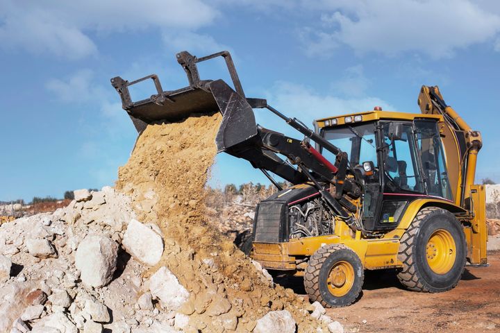 Yellow backhoe dumping a load of dirt onto a pile of rocks against a blue sky.