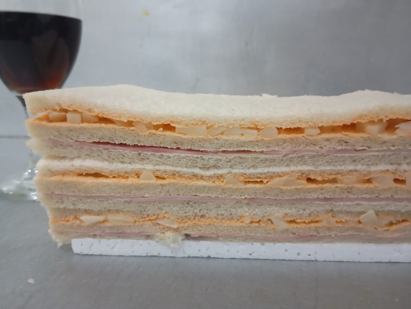 Corte transversal de un sándwich de varias capas, relleno de jamón y queso, junto a un vaso de líquido rojo.