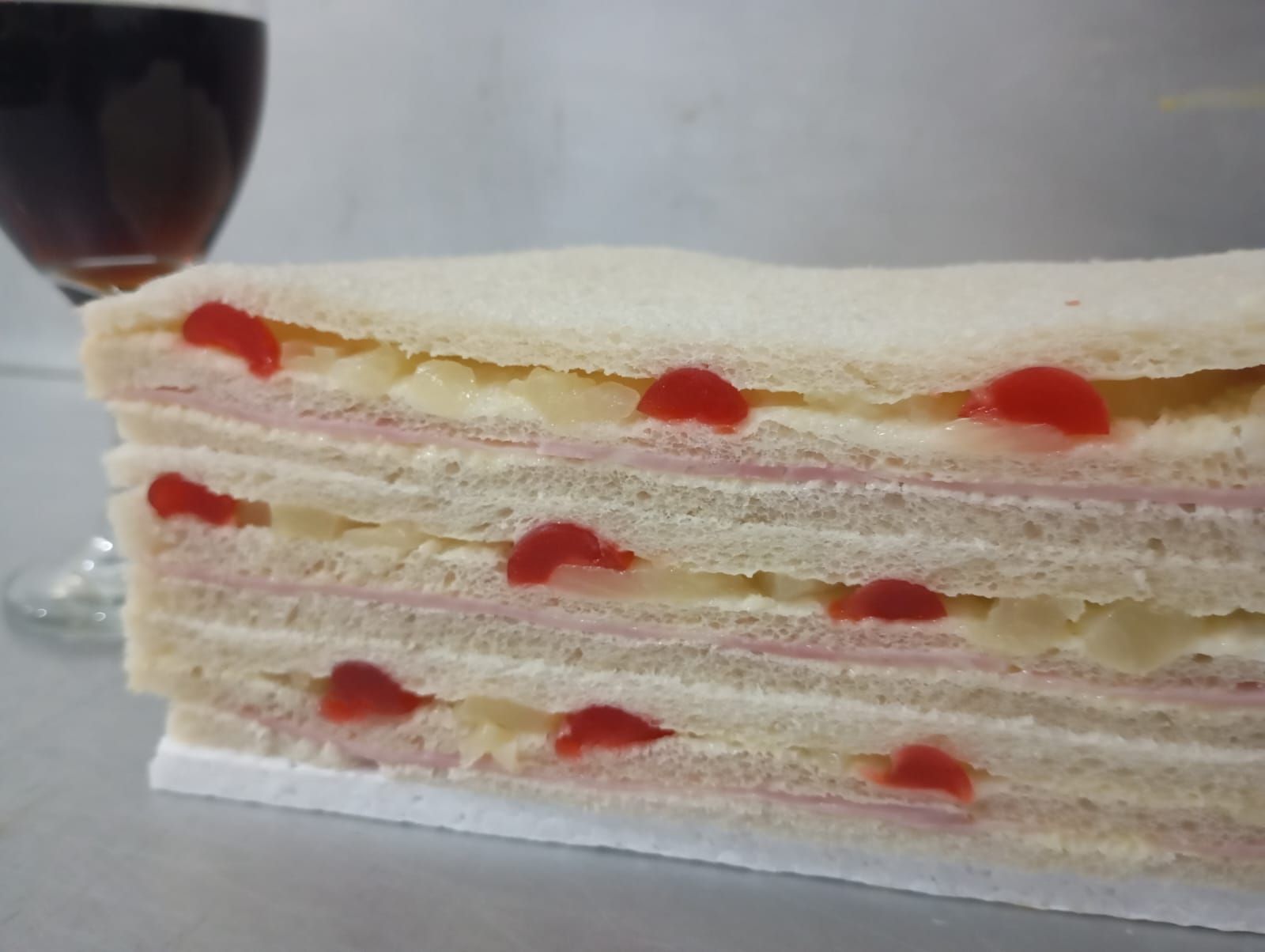 Sándwich de capas con jamón, queso y cerezas rojas, sobre una superficie blanca, con un vaso de líquido oscuro en el fondo.