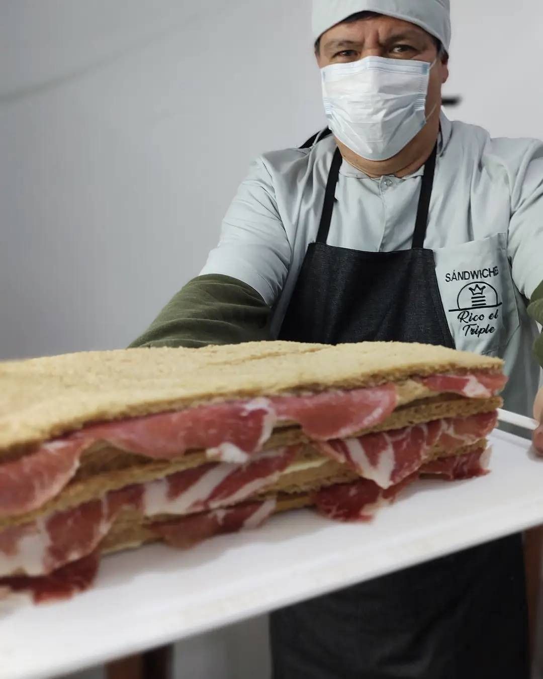 Hombre sosteniendo un sándwich grande con varias capas de jamón. Lleva mascarilla y delantal.