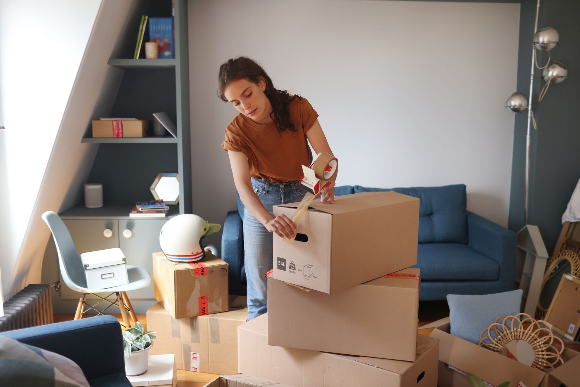 A woman sealing cardboard boxes in a living room filled with packed items