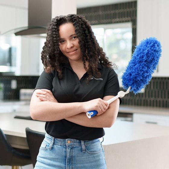 Woman in a black shirt and jeans holding a blue duster in a modern kitchen, arms crossed, smiling.