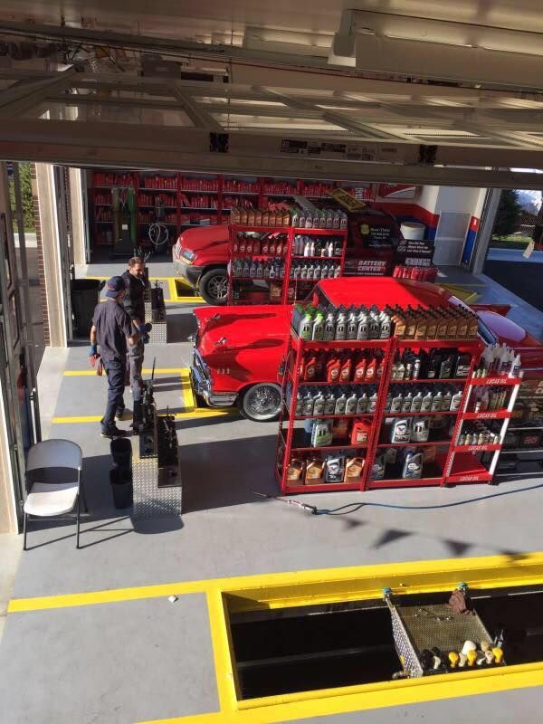 Two men near red classic cars with oil bottles in a service bay.