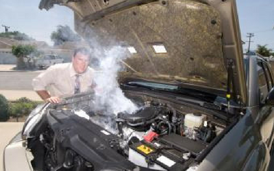A man looks into the engine bay of a car emitting thick white smoke, parked on a residential street.