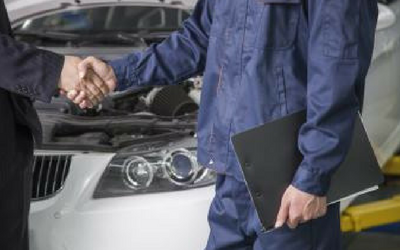 A person in a blue uniform holds a clipboard and shakes hands with a customer in a garage with an open car hood.