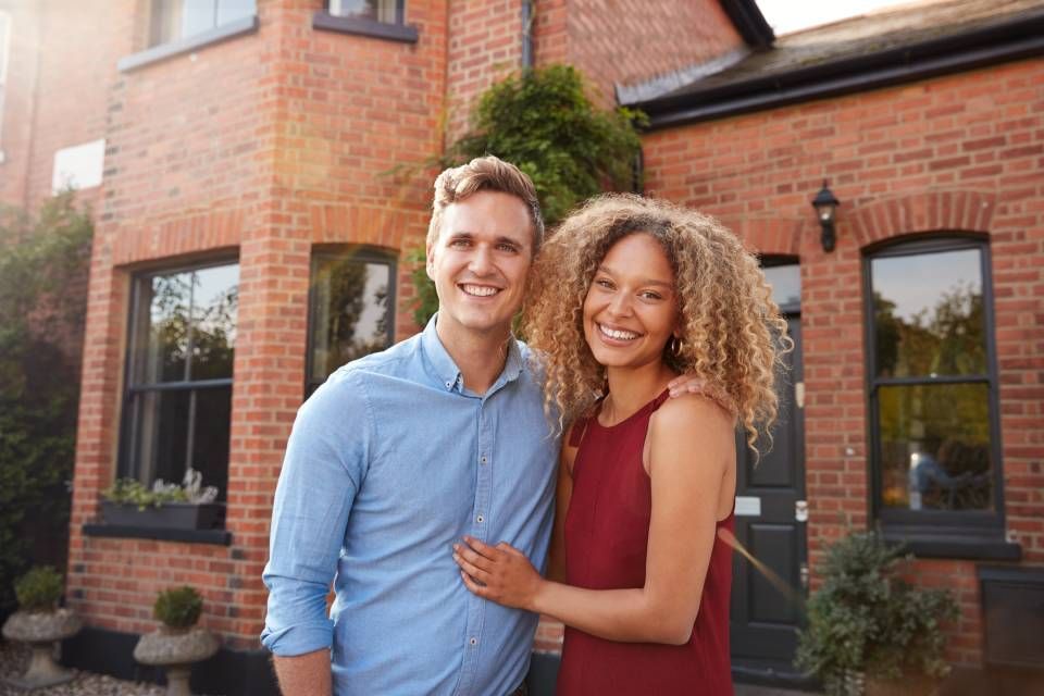 A smiling couple stands in front of a brick house, embracing each other warmly.
