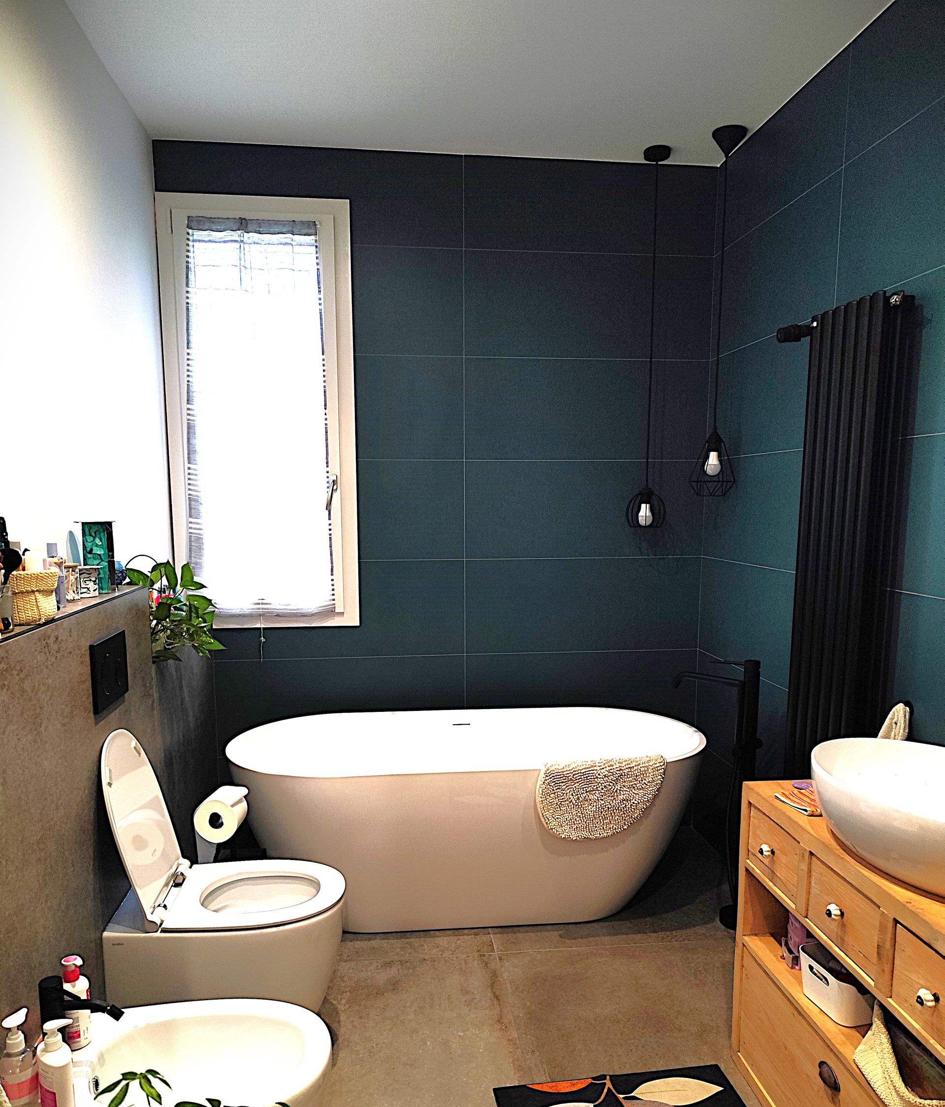 Bagno con vasca bianca indipendente, pareti color verde acqua, finestra, WC, lavandino e mobiletto in legno.