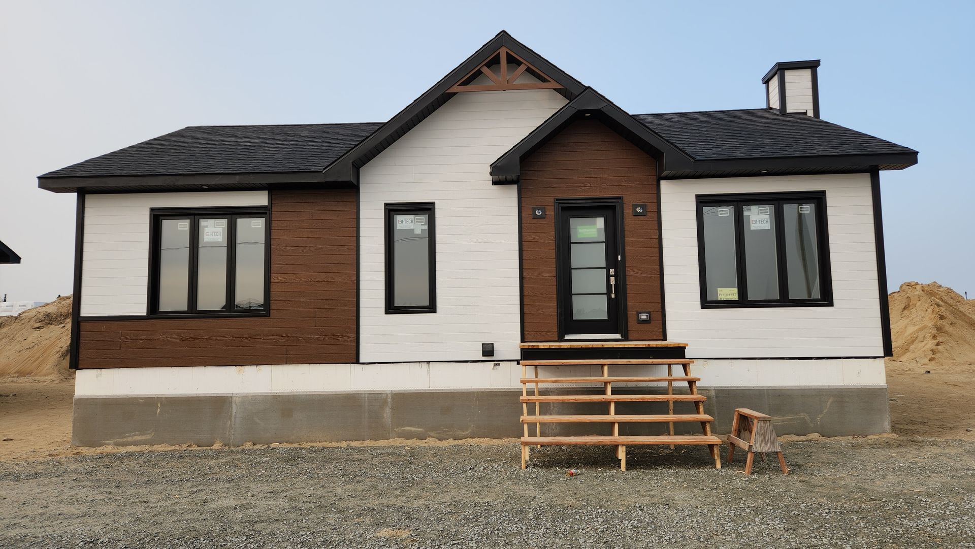 Une petite maison au toit noir se trouve au sommet d’un terrain de gravier.