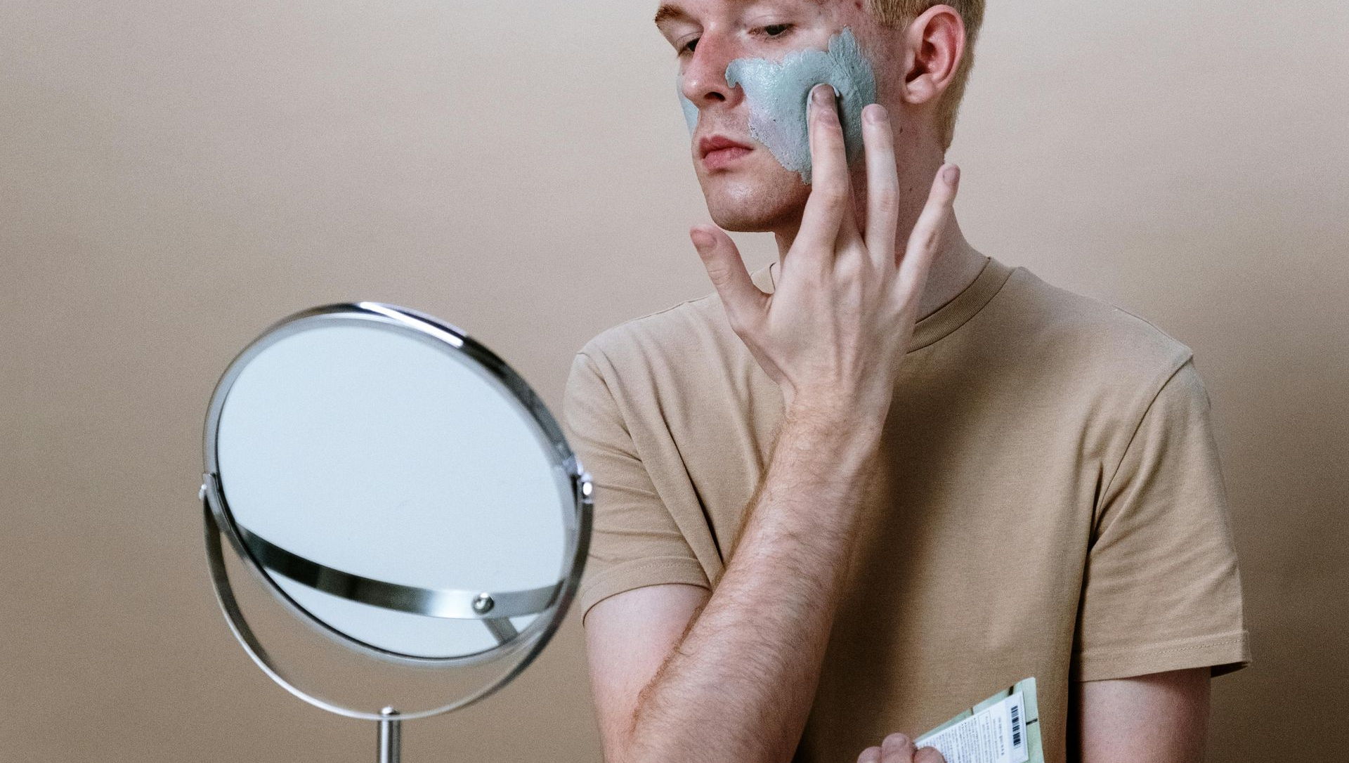 A person applies a facial mask, looking at a round mirror. Beige background, light skin, light shirt.