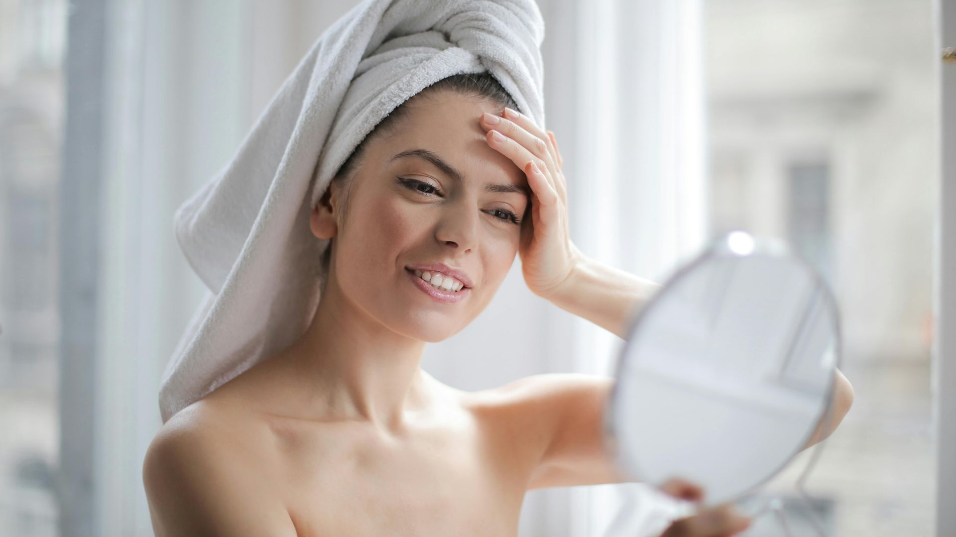 Woman with towel on head, looking in a hand mirror, touching her forehead, indoors near a window.