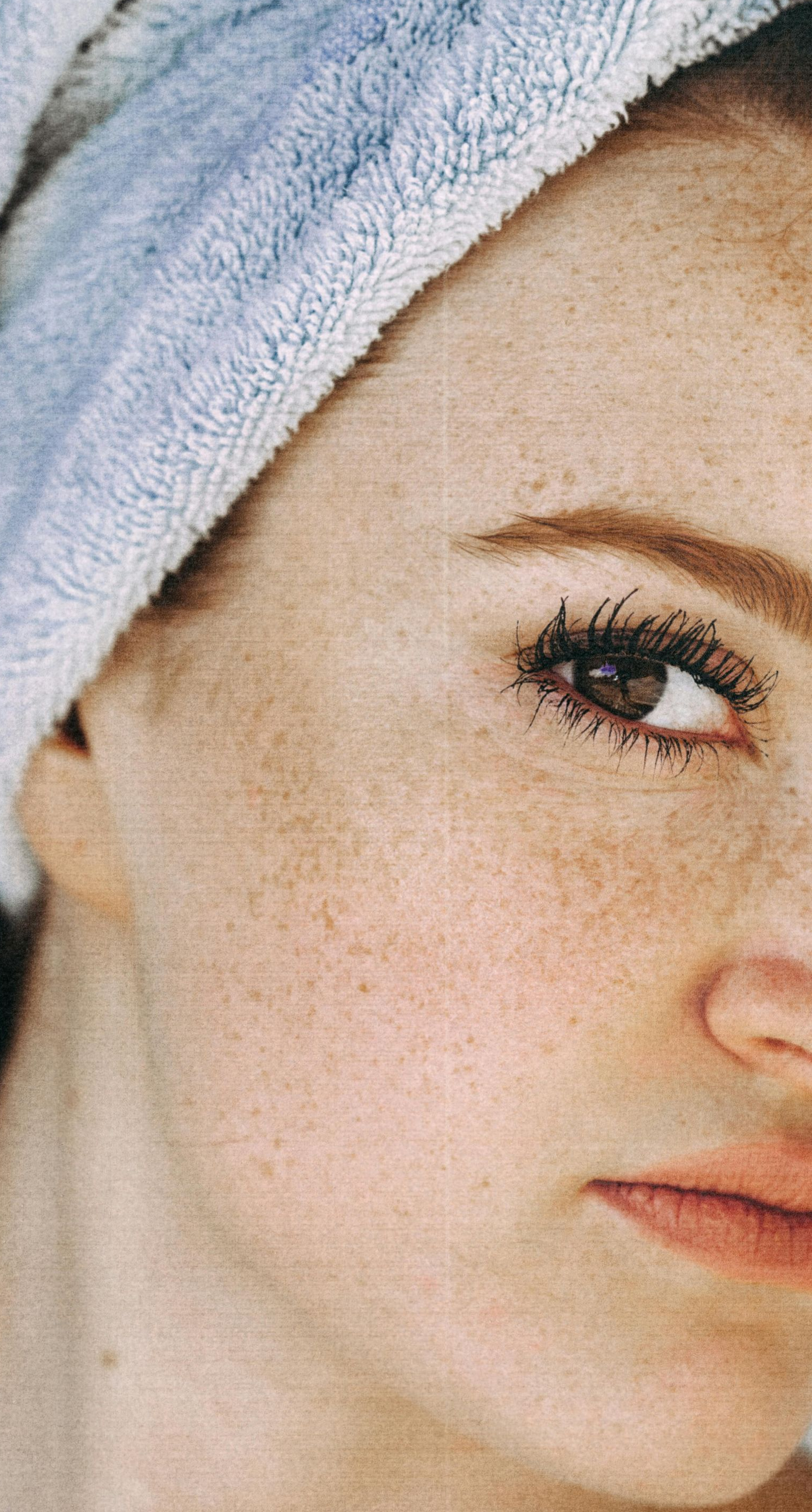 Woman with freckled skin, wrapped in a light blue towel, looking down.