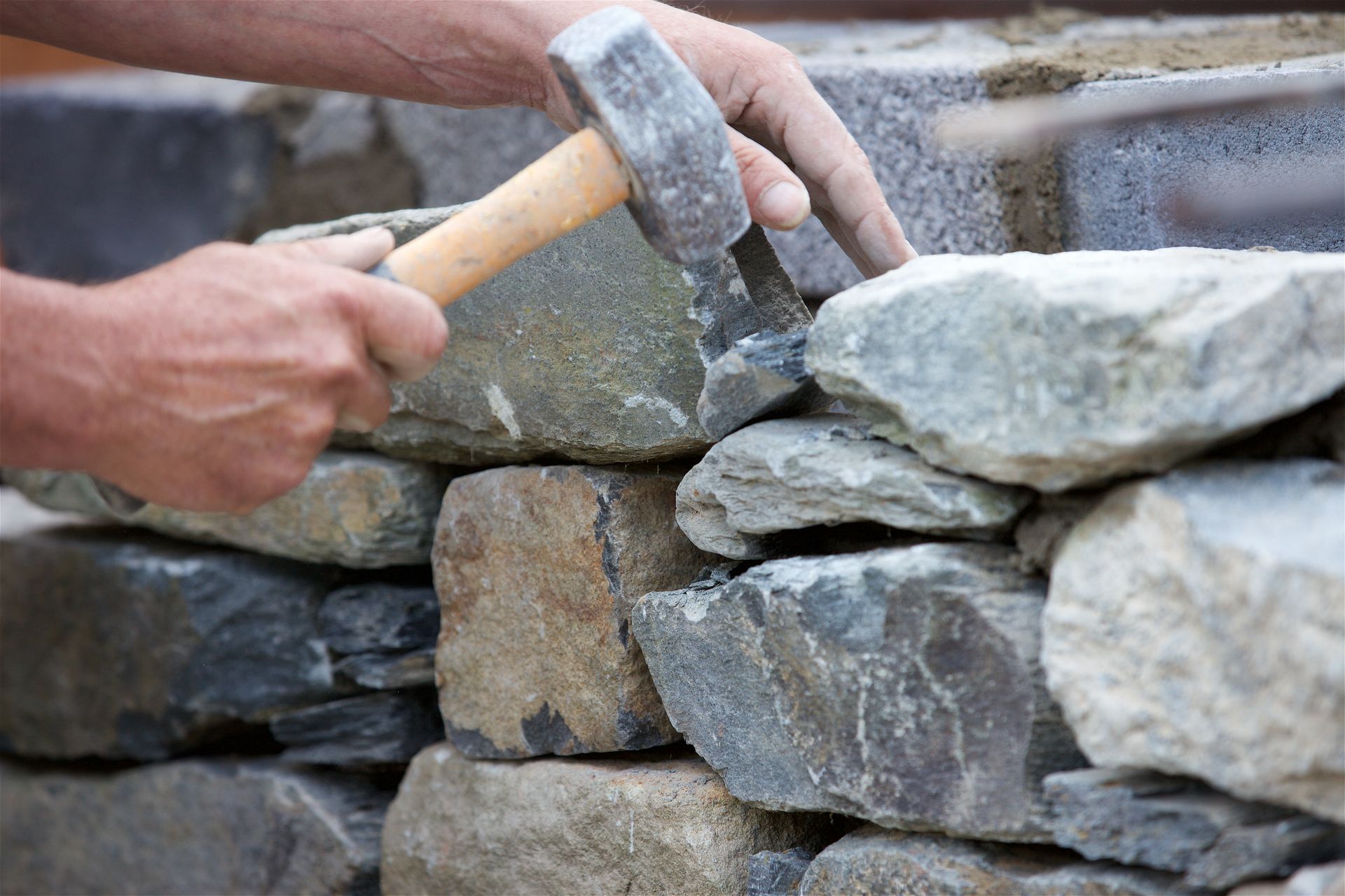 Hands using a hammer to shape stone while building a wall.