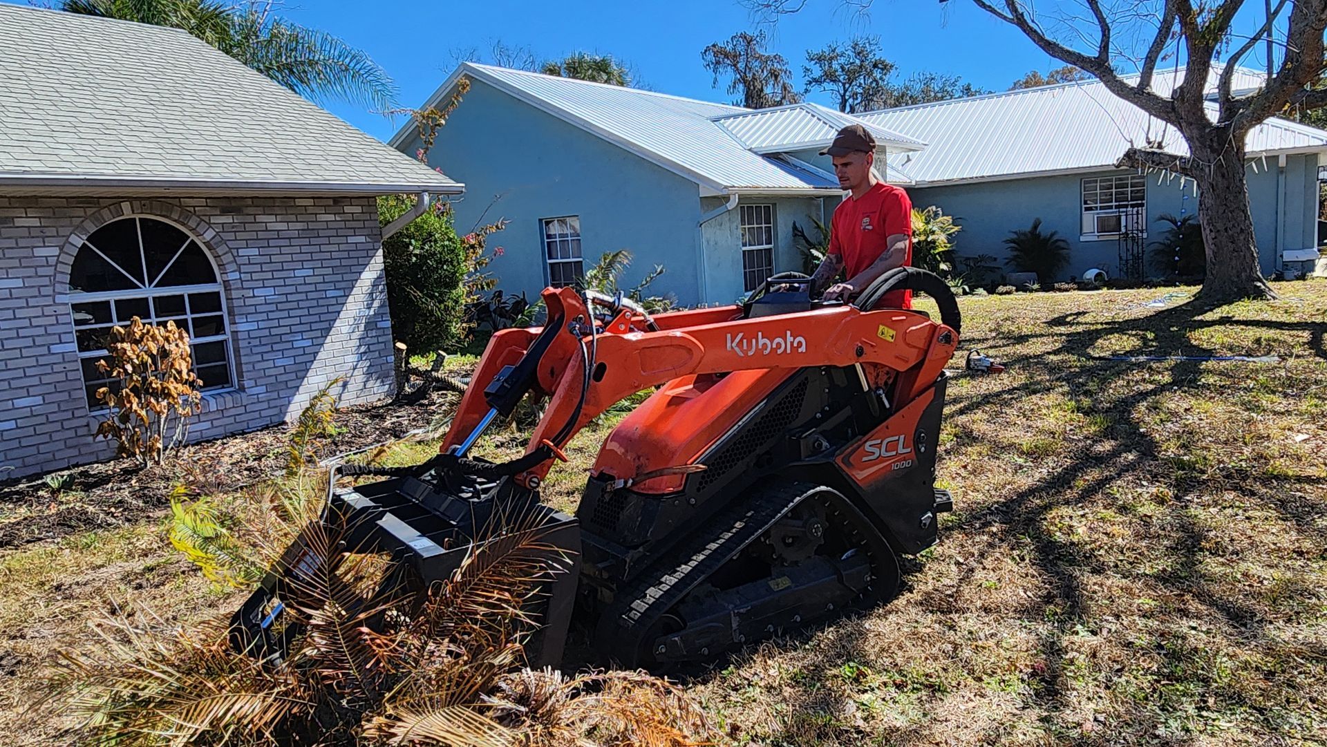 Person on an orange Kubota tractor clearing debris in a yard beside a house with trees and a blue home in the background