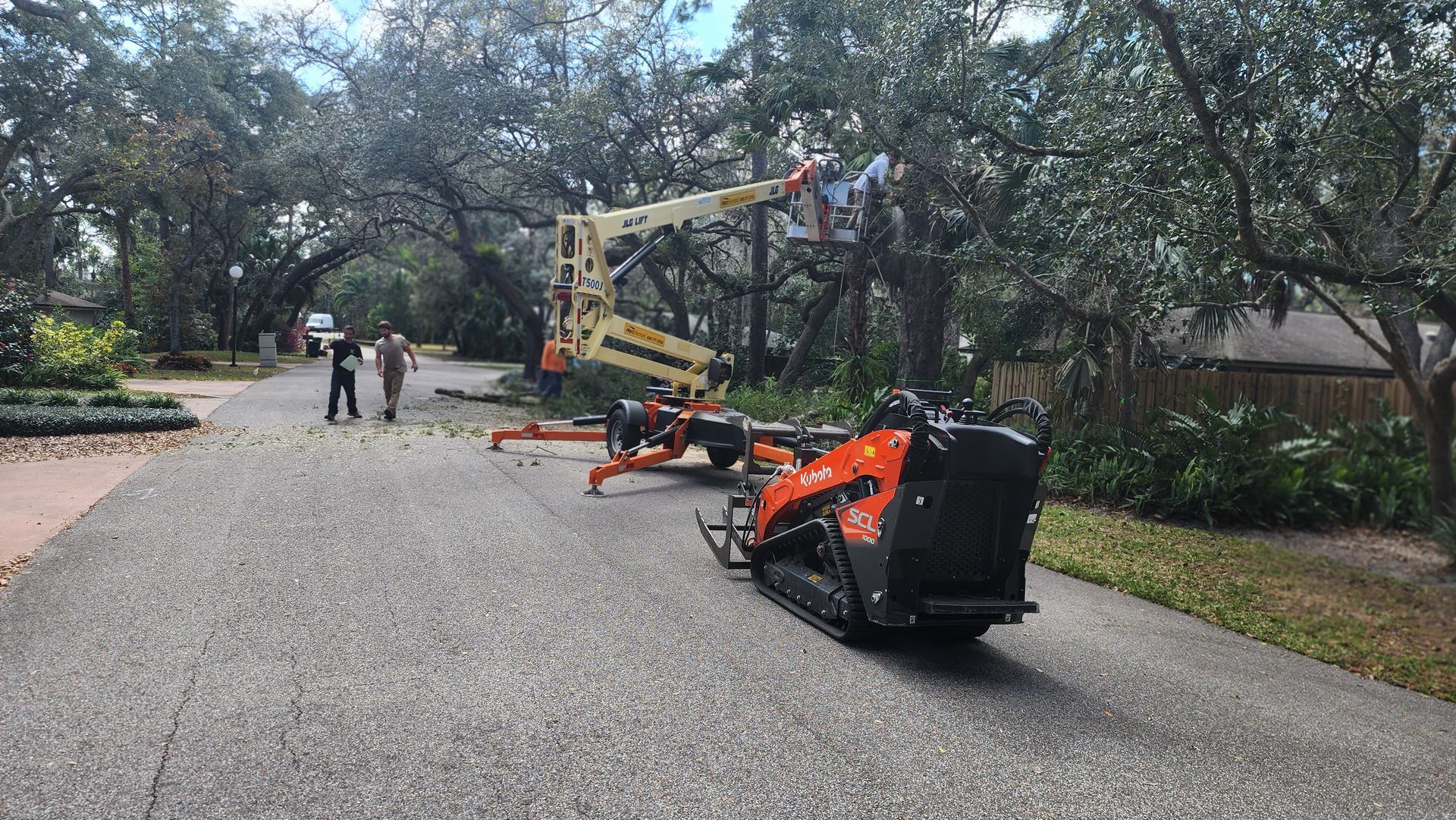Orange excavator and boom lift on a gravel road under trees, with two people walking in the distance.