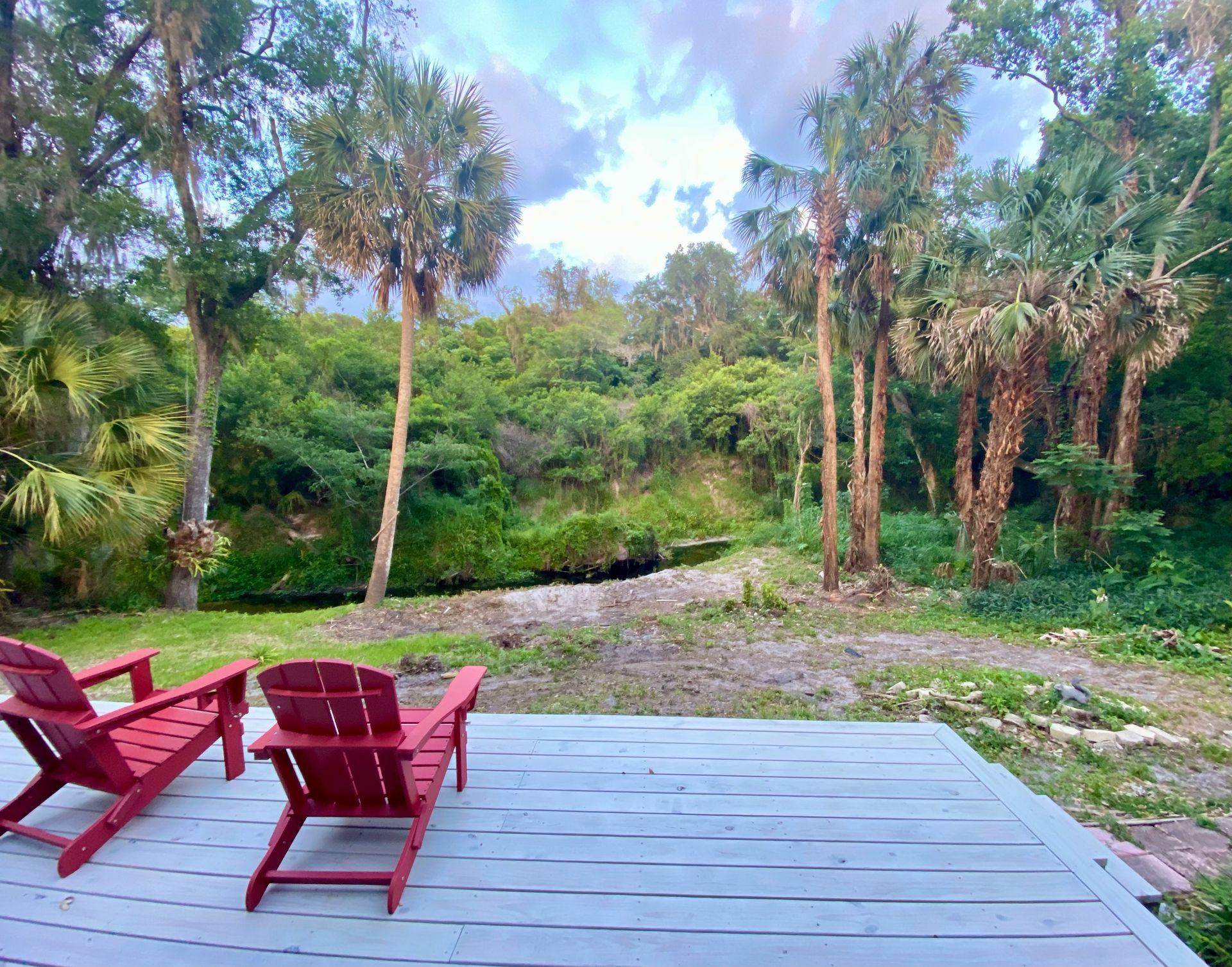 Wooden deck with red chairs overlooking a lush green garden and trees under a blue sky.