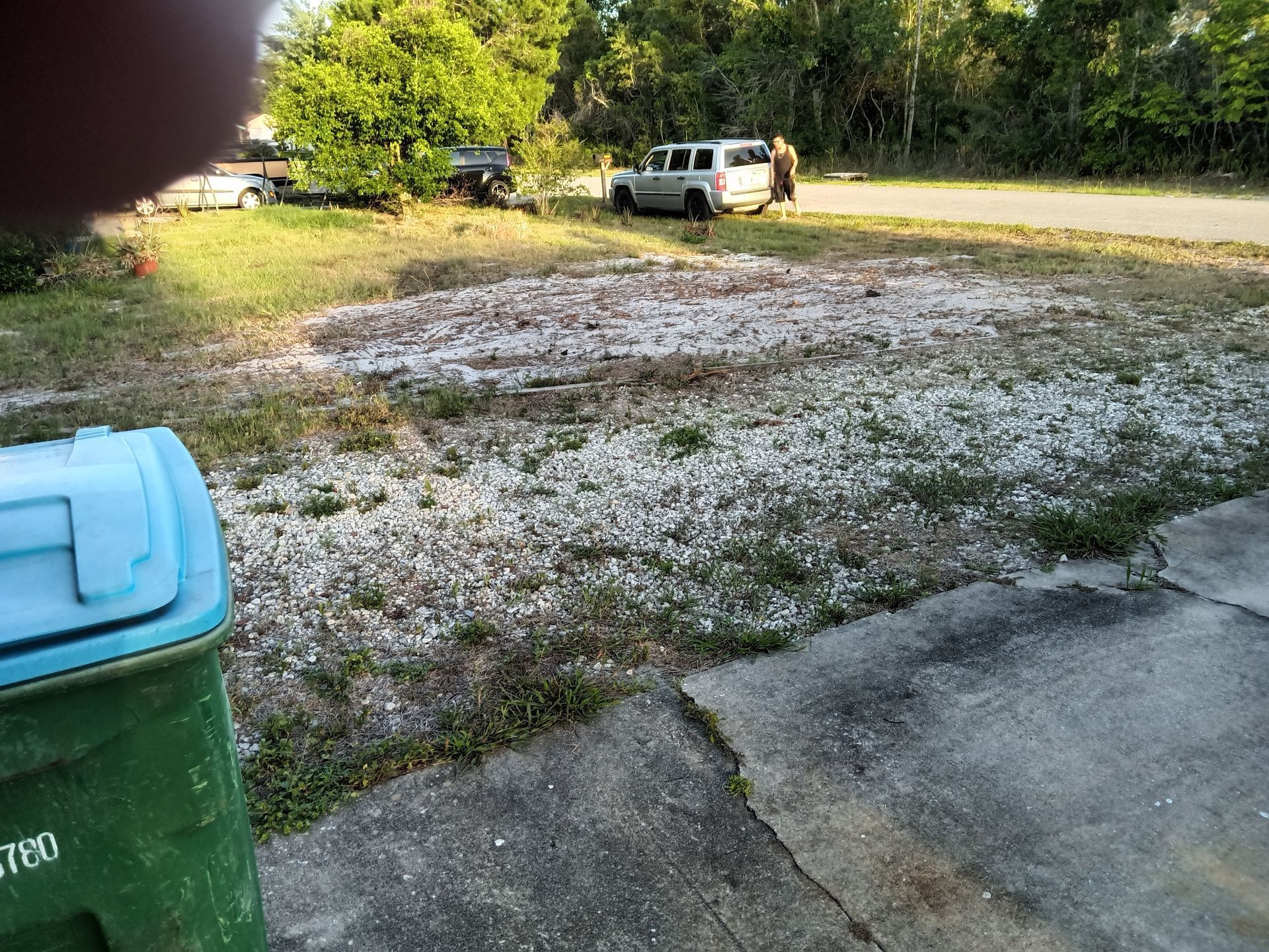 Gravel driveway and grassy yard with a white truck near trees, viewed from a concrete porch.