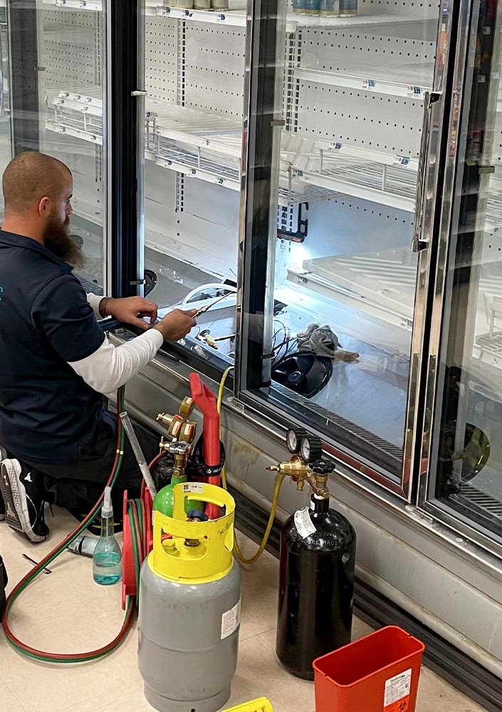 A technician repairs a refrigerated display case in an empty supermarket aisle. They use welding equipment.