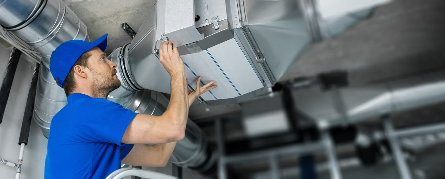 A technician in a blue uniform working on an HVAC system, installing a filter.