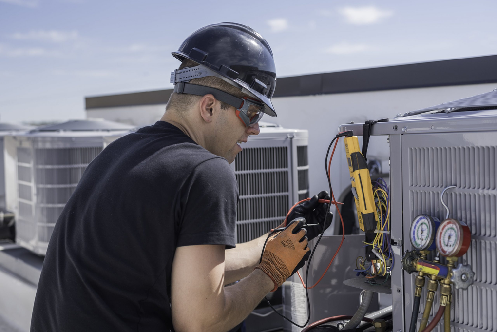 HVAC technician on a rooftop, working on an air conditioning unit with a multimeter and gauges.
