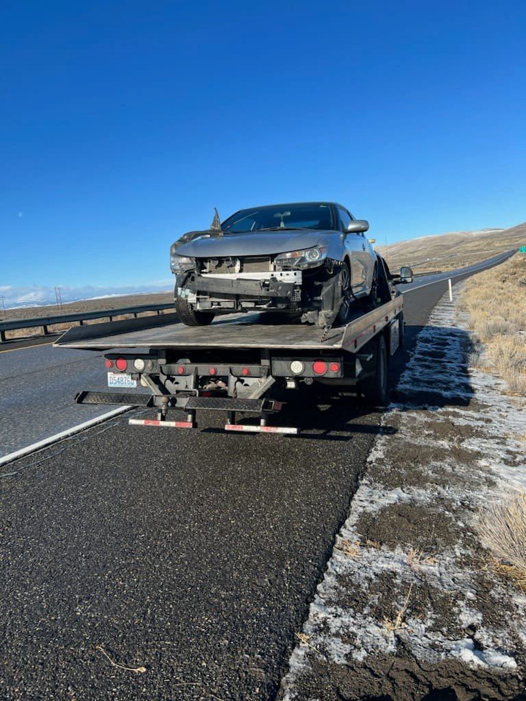 Damaged silver car loaded onto a flatbed tow truck on a rural road under a blue sky.