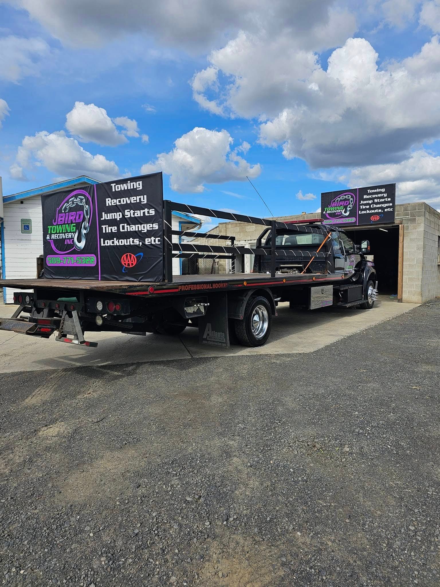 Black flatbed tow truck with business logos, parked outside on a gravel lot under a cloudy sky.