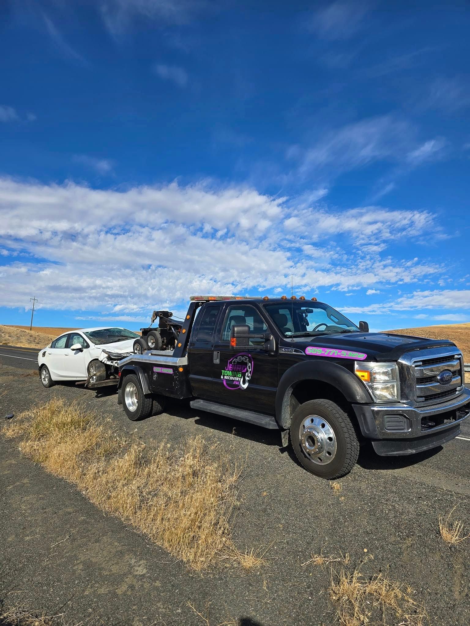Tow truck towing a white car on a road under a blue sky with clouds.