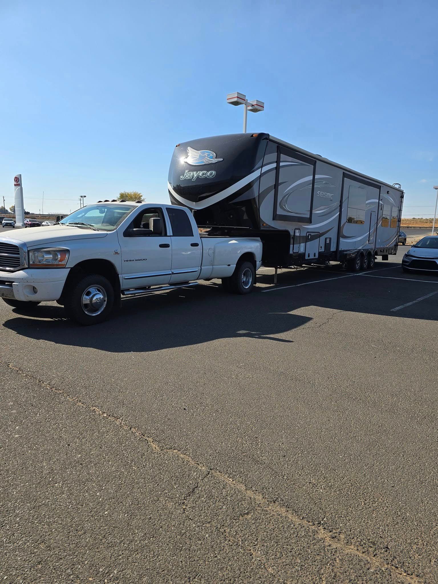 White pickup truck towing a large, black and grey Jayco fifth wheel trailer on a gravel lot under a blue sky.
