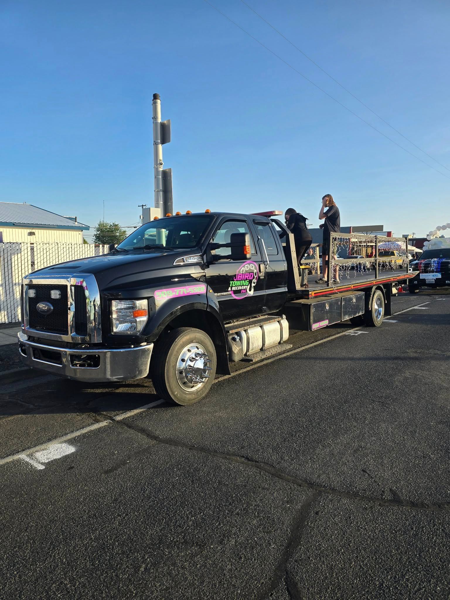 Black flatbed tow truck parked on asphalt on a sunny day. A person stands on the bed.